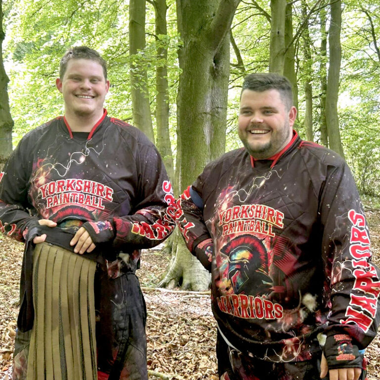 Two men in Yorkshire Paintball Warriors jerseys smiling in a wooded forest setting.